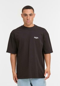 Mand med fregner iført en løs sort kortærmet skjorte med lille hvidt logo, lyseblå jeans, stående foran en ensfarvet hvid baggrund.