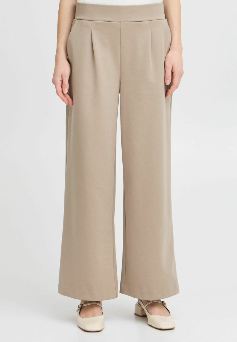 Pantalons larges beiges en matériau lisse, dotés de plis avant et d'une taille haute. Associés à des chaussures de couleur claire.