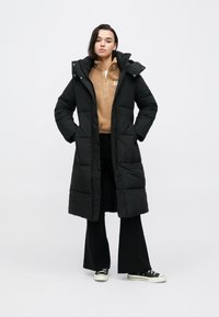 Fekete pufidzseki hosszú fazonban, kapucnival és nagy zsebekkel. Bézs polár felső és fekete nadrág fölött viselve, tornacipővel párosítva.