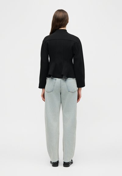 Γυναίκα με μακριά καστανά μαλλιά που φοράει μαύρο μακρυμάνικο μπουφάν και ανοιχτό μπλε φαρδιά τζιν, στέκεται γυρισμένη πλάτη σε λευκό φόντο.