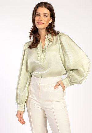 Lichtgroene satijnen blouse met gerimpelde kraag en knopenlijst, met pofmouwen. Gepaard met hoge crèmekleurige broeken.
