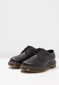 Des chaussures brogues en cuir noir avec des détails perforés, bout rond et couture jaune contrastante sur une semelle épaisse et translucide.