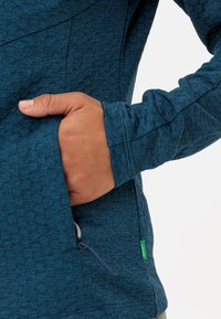 Giacca turchese con motivo, con una mano in una tasca laterale, presenta un cordoncino e un'etichetta verde sul bordo. Dettaglio di tessuto testurizzato visibile.