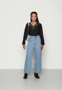 Zwarte lange mouwen top met kantdetail bij de decolletés, gecombineerd met lichtblauwe high-waisted wijde jeans en zwarte laarzen.