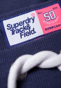 Nahaufnahme eines marineblauen Stoffetiketts mit der Aufschrift "Superdry Track & Field" und "SD Athletic Dept." auf rotem Hintergrund sowie einer weißen Kordel.