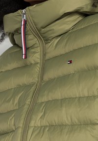 Doudoune vert olive avec un col haut, des coutures horizontales et une fermeture éclair métallique dotée d'une tirette rouge, blanche et bleue. Logo affiché.