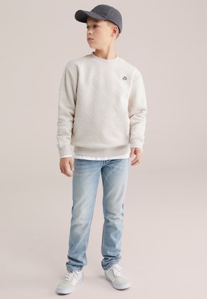 Hellbeiger Pullover mit strukturiertem Rautenmuster, getragen über einem weißen Hemd, kombiniert mit hellblauen Jeans und grauer Mütze, ergänzt durch weiße Turnschuhe.