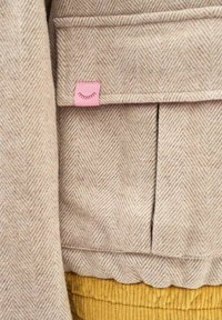 Beežika kala luu mustriga kangas, millel on väike roosa silt, millel on suletud ripsmekujundus, ja allpool sinepikollane soonikkangas.