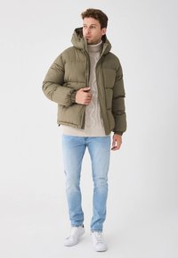 Oliwkowa pikowana kurtka z kapturem, ściągaczami przy rękawach, zapinana na zamek. Nałożona na kremowy sweter w warkocze oraz jasnoniebieskie jeansy z białymi sneakersami.