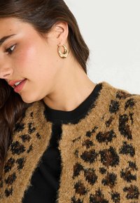 Cárdigan de leopardo en tonos marrones con textura suave, camiseta negra debajo y pendientes de aro dorados. Énfasis en el escote y el perfil facial.