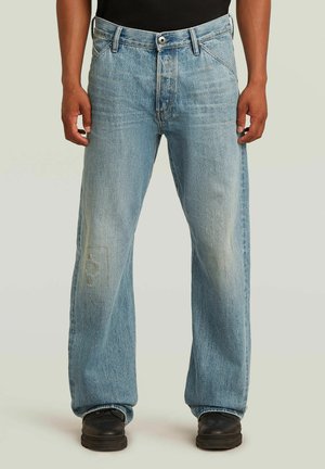 Jeans a gamba dritta di colore azzurro chiaro con lievi effetti consumati, indossati con scarpe nere e una camicia nera su uno sfondo semplice.