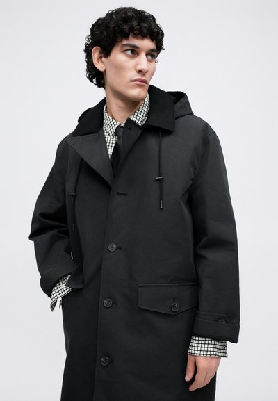 Man wearing a long black coat with a hood over a patterned shirt and black tie, looking to his left with one hand in his pocket.