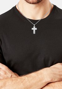 Mann trägt ein schwarzes T-Shirt und eine silberne Kreuzanhänger-Halskette, die Arme vor der Brust verschränkt.