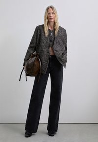 Blazer texturizado gris sobre una blusa negra, combinado con jeans negros de pierna ancha. Sosteniendo un bolso de cuero marrón con cordón, calzado con zapatos negros.