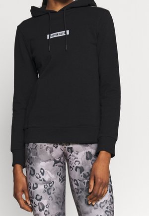 Personne portant un sweat à capuche Calvin Klein noir avec logo et des leggings gris à imprimé léopard, debout devant un fond clair uni.