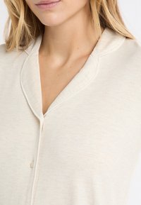 Jasnobeżowy kardigan o miękkiej fakturze, z dekoltem w serek, subtelnym przeszyciem i zapinany na jeden guzik.