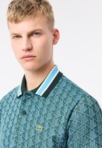 Grönmönstrad polotröja med geometrisk design, som har en blå och vit randig krage samt en liten broderad logo på bröstet.