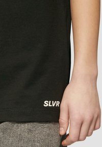 Nahaufnahme einer Person, deren Hand neben einem schwarzen Shirt liegt, auf dem "SLVR" am Saum gedruckt ist, sowie grauen strukturierten Hosen.