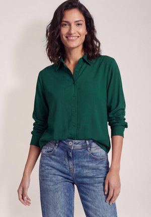 Mujer con cabello oscuro hasta los hombros, con una camisa verde de manga larga abotonada y vaqueros azules, de pie frente a un fondo sencillo.