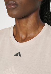 Jasnoróżowa sportowa koszulka z okrągłym dekoltem i czarnym logo Adidas po lewej górnej stronie, wykonana z gładkiego materiału o luźnym kroju.