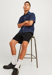 Short-sleeve denim shirt in dark blue, paired with black shorts. Model sits on a metal stool, wearing white socks and black shoes.