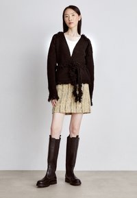 Brązowy sweter z fakturą, z postrzępionymi krawędziami, zestawiony z plisowaną, beżową mini spódnicą i brązowymi skórzanymi kozakiami do kolan.