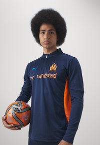 Tamsiai mėlyna sportinė marškinė su oranžinėmis akcentais, turinti stovintį apykaklę ir logotipą. Žaidėjas laiko daugiakampį, įvairiaspalvį futbolo kamuolį.