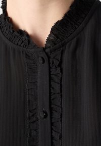 Blusa nera con colletto arricciato, righe verticali e una sottile trama. Chiusura con bottoni neri sulla parte frontale.