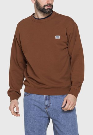 Uomo che indossa una felpa marrone a maniche lunghe e jeans blu, con un piccolo logo rettangolare sul lato sinistro del petto.