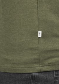 Olive green t-shirt with a soft texture, featuring a white tag with an exclamation mark at the hem. Black denim visible below.
