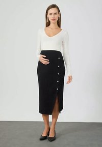 Femeie îmbrăcată cu un top alb cu mânecă lungă și fustă neagră cu nasturi, ținându-se de burtă, stând pe un podea gri, pe un fundal gri simplu.