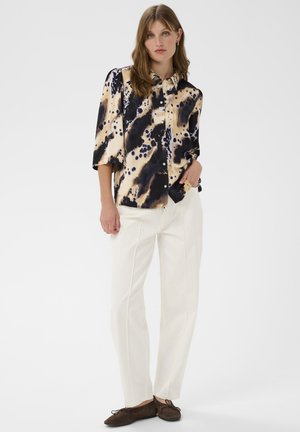 Ženska oblečena v črno-bež majico s potiskom, z belimi ravnimi hlačami in rjavimi ravnimi čevlji, stoji z eno roko v žepu.