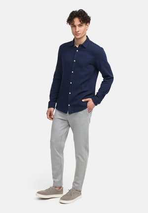 Navyblaues Langarm-Hemd mit Knopfleiste aus strukturiertem Stoff, kombiniert mit hellgrauen, schmal geschnittenen Jogginghosen und grauen Schnürschuhen.