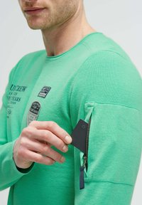 Grünes, strukturiertes Langarmshirt mit einer Reißverschlusstasche am Ärmel; enthält Logo-Patches und einen runden Ausschnitt.