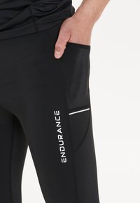 Svarta tights för träning med sidficka, prydda med en vit logotyp "ENDURANCE" och en smal vit rand för detaljer. Slät, sträckbar tyg.