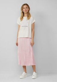T-shirt crème avec un motif floral et du texte, associé à une jupe midi plissée rose et des baskets blanches. Design simple, texture douce.