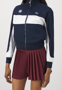 Kvinna iförd en marinblå, vit och vinröd zip-up sportjacka med Roland Garros och Lacoste-logotyper samt en vinröd plisserad tenniskjol.