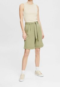 Shorts vert olive avec une taille ceinturée, un design plissé et des ourlets roulés, associés à un haut sans manches beige et des baskets claires.