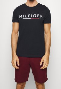 Czarny bawełniany t-shirt z białym i czerwonym logo "HILFIGER" na piersi. Krótkie rękawy i okrągły dekolt. Połączony z burgundowymi szortami.