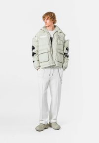 Chaqueta de plumón gris claro con bordado floral negro, cuatro bolsillos frontales, puños ajustables, combinada con pantalones blancos y zapatos grises texturizados.