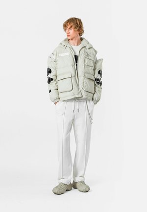 Chaqueta de plumón gris claro con bordado floral negro, cuatro bolsillos frontales, puños ajustables, combinada con pantalones blancos y zapatos grises texturizados.