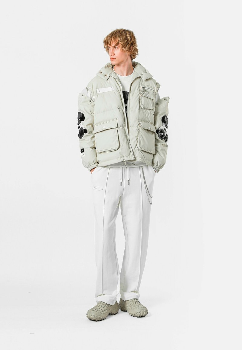 Chaqueta de plumón gris claro con bordado floral negro, cuatro bolsillos frontales, puños ajustables, combinada con pantalones blancos y zapatos grises texturizados.