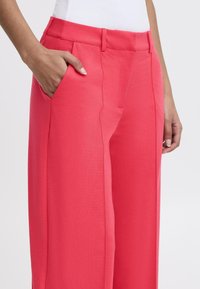 Pantalons tailleur roses avec une coupe décontractée, poches latérales et texture lisse ; couture visible le long de l'avant et taille mi-haute.