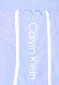 Pantalones cortos deportivos de color azul claro con rayas verticales blancas y paneles de malla. "Calvin Klein" impreso verticalmente en blanco en el lateral.