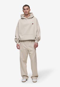 Beige hoodie with a front pocket, drawstring hood, and embroidered logo. Paired with matching beige pants and white sneakers.