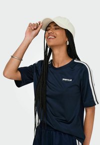 Marineblaues Sport-T-Shirt mit weißen Seitenstreifen, aus glattem Material. Eine hellfarbige Mütze wird getragen, mit geflochtenem Haar und einem silbernen Armreifen sichtbar.