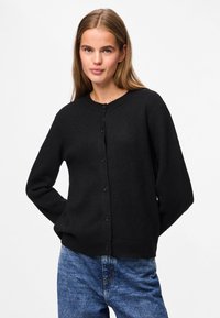 Cardigan noir en tricot avec encolure ronde, manches longues et fermeture à boutons, doté d'une texture douce et d'un ourlet côtelé. Porté avec un jean bleu.