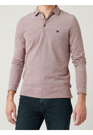 Homme portant un polo manches longues rose poudré avec un tissu texturé subtil et un jean foncé, debout devant un fond clair uni.