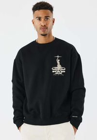 Jongeman met korte krullen, gekleed in een zwart sweatshirt met een beige afbeelding van het Vrijheidsbeeld en witte broek, met de handen in de zakken, op een neutrale achtergrond.