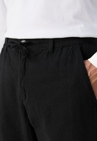 Zwarte, lichte broek met een elastische tailleband, trekkoord en zijzakken. De stof heeft een gladde textuur.